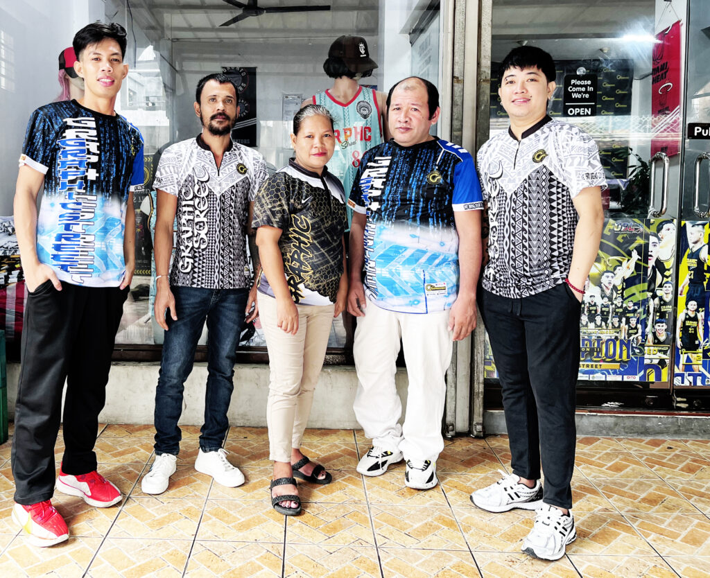 About Five Graphicstreet production staff members, three men and two women, standing outside a storefront, all wearing different designs of Graphicstreet full sublimation jerseys. The jerseys feature diverse patterns in black, white, blue, and gold.